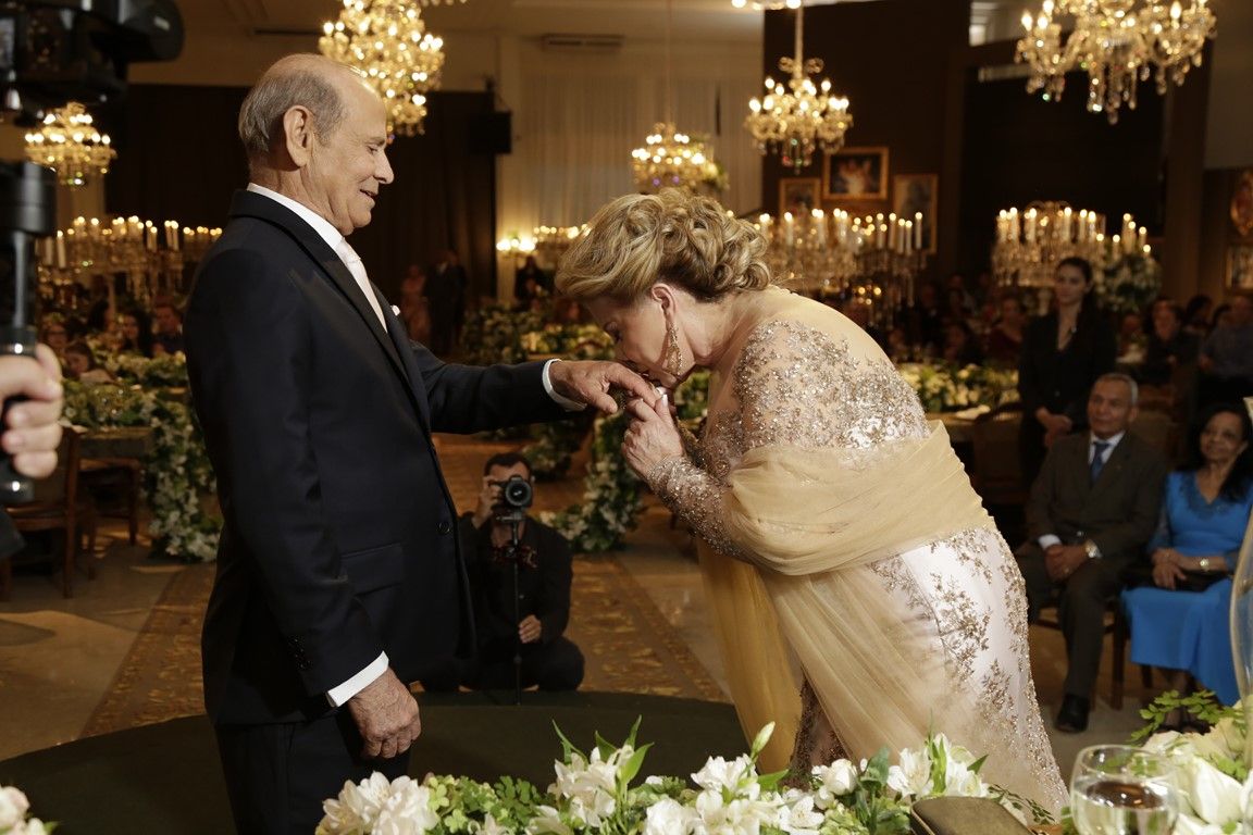 Foto de casal comemorando as bodas do casamento. A mulher está de dourado e beija a mão do marido.