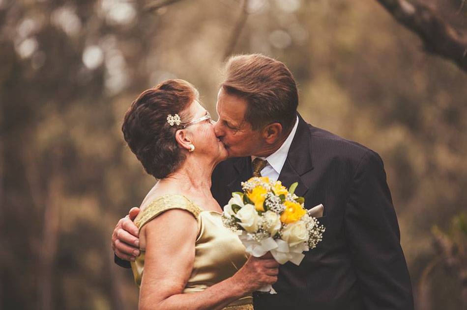 Foto de casal comemorando as bodas de 50 anos de casamento. A mulher está vestindo um vestido dourado, segurando um buquê de flores e beijando seu marido