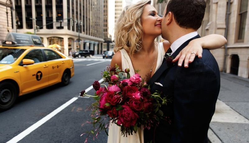 Foto colorida de casal se acariciando. Eles estão na rua e a mulher está com um buquê de flores na mão.