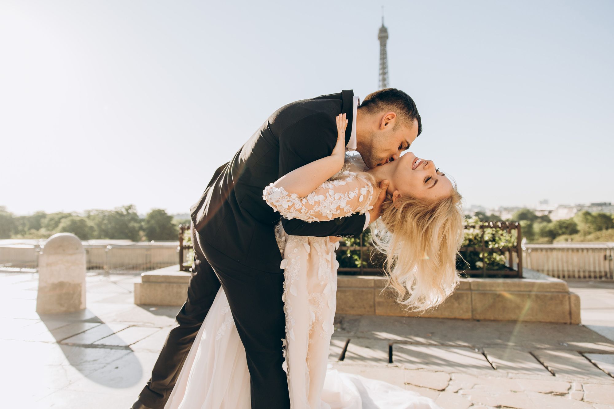 Foto colorida de casal em frente à torre eiffel, em paris. O noivo está debruçando a mulher sobre seus braços es e beijando seu pescoço. Ambos estão vestindo trajes de noivos.