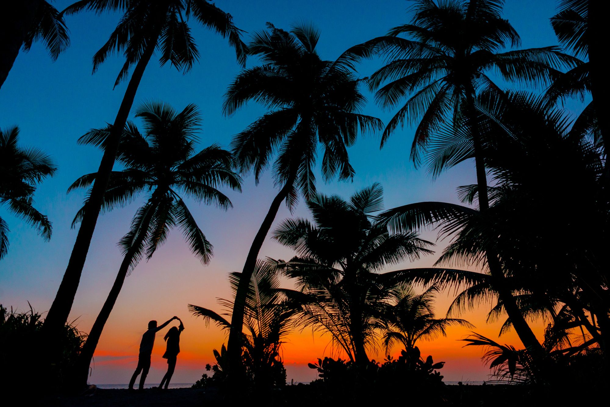 Foto colorida de por do sol na praia, com casal dançando na areia.