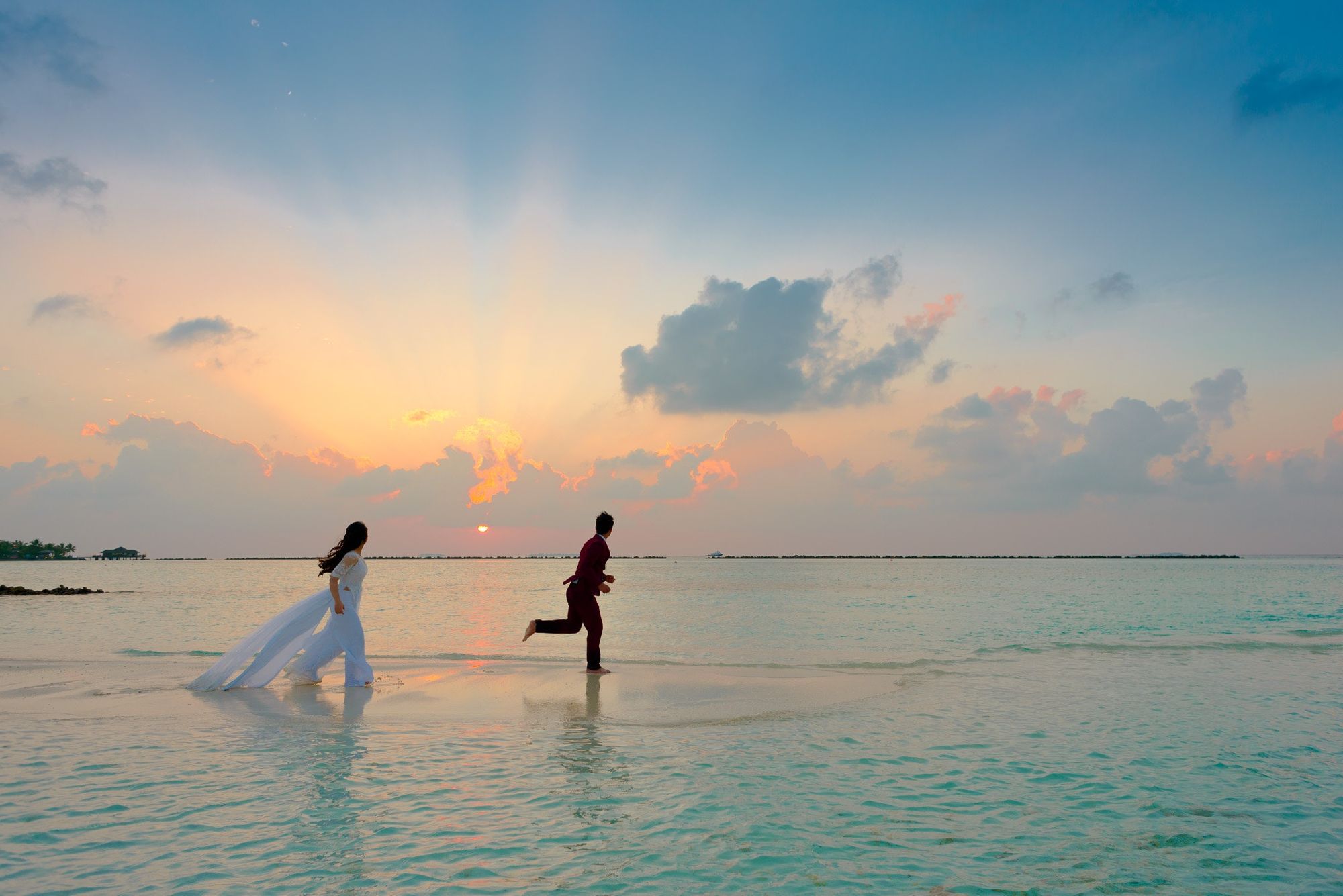 Foto colorida de casal correndo em praia. Eles estão vestidos de noivos e o dia está acabando.