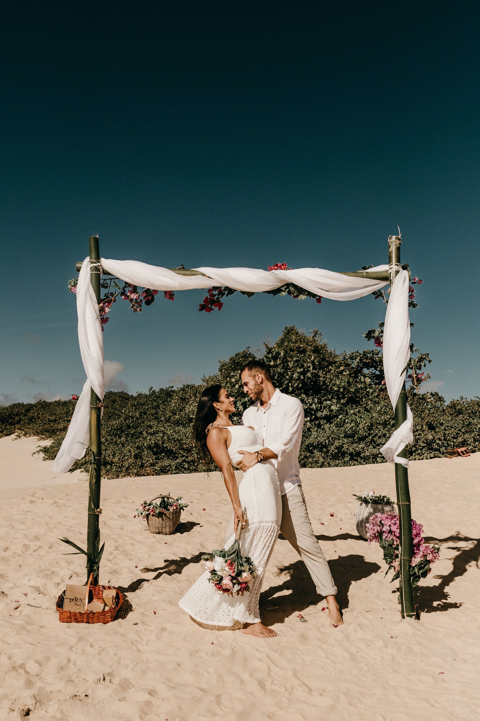 Foto colorida de casal recém casados na praia. O noivo está segurando a cintura da mulher e olhando fixo em seus olhos.