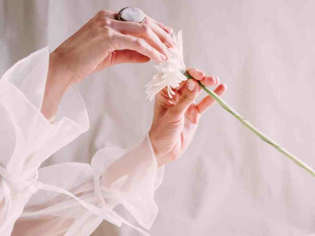 As mãos de uma mulher segurando uma flor branca. As mangas de seu vestido são bufantes, brancas e com transparência.