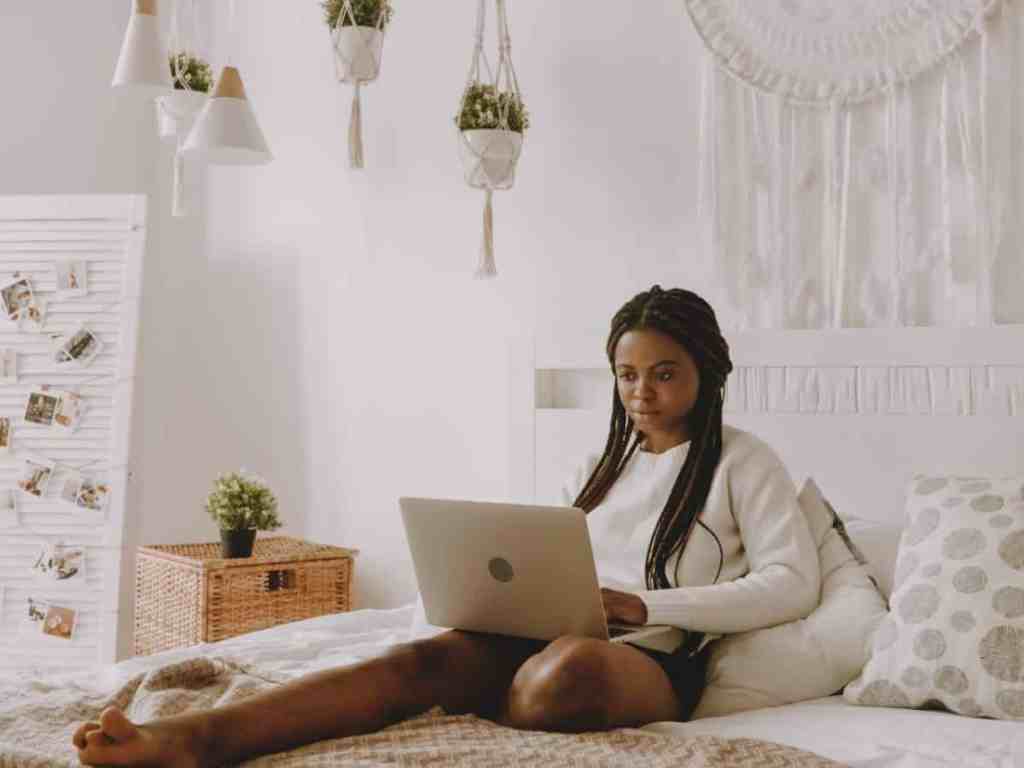 Mulher está sentada encostada na cama com um notebook no colo, ela usa blusa branca de manga longa e short curto preto.