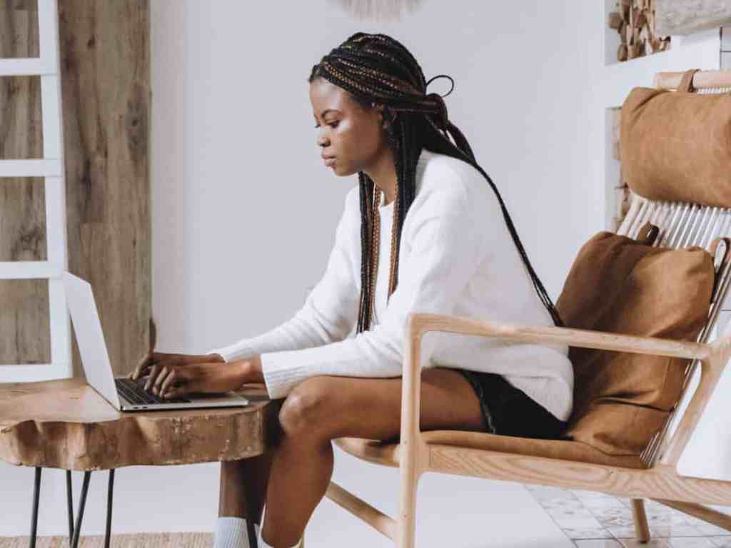 Mulher de tranças box braid, usa blusa de manga longa branca e short preto, está sentada em uma cadeira em casa mexendo no notebook que está sobre uma mesinha.