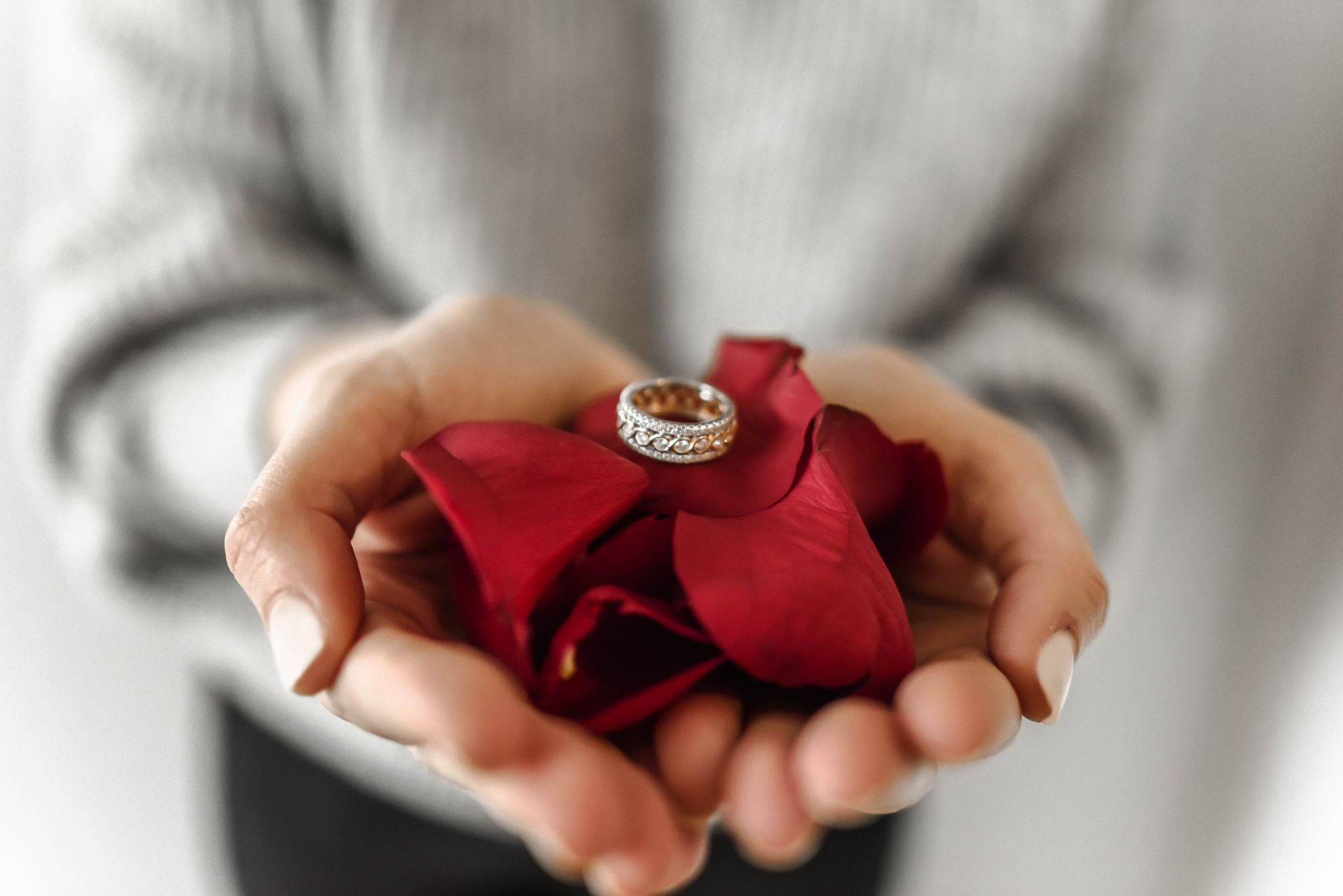 Foto colorida de noiva segurando a aliança em cima de pétalas de rosas vemelhas. O fundo está desfocado.