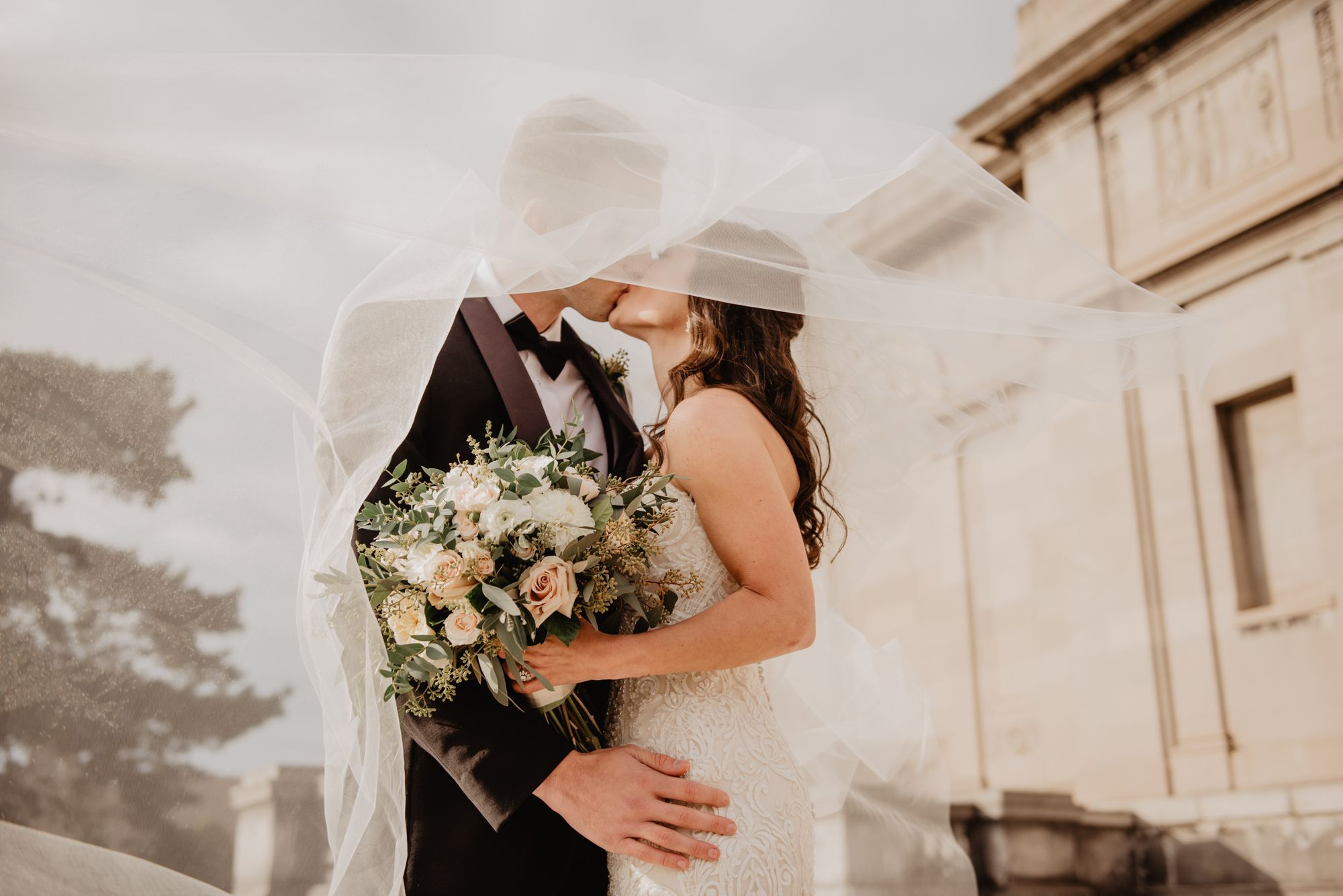 Foto colorida de noivos se beijando em baixo de véu. A noiva segura um buquê de flores.