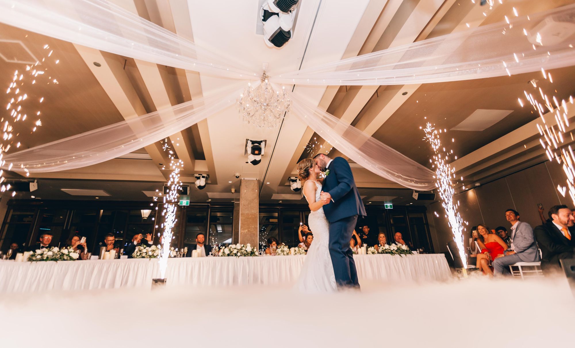 Foto colorida de noivos dançando e se beijando no meio do salão.