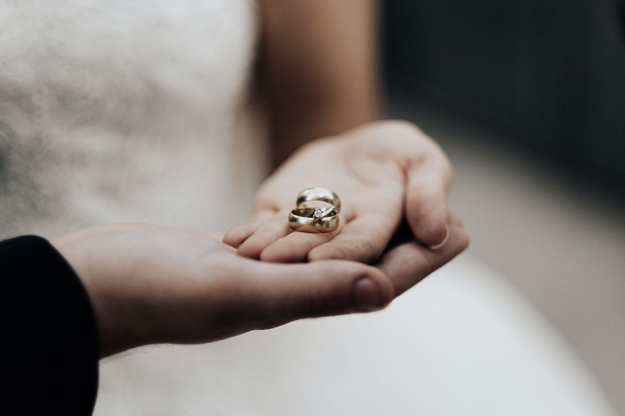 Foto colorida de noivos segurando as alianças nas palmas dão mãos. A mão do noivo está embaixo da mão da noiva.