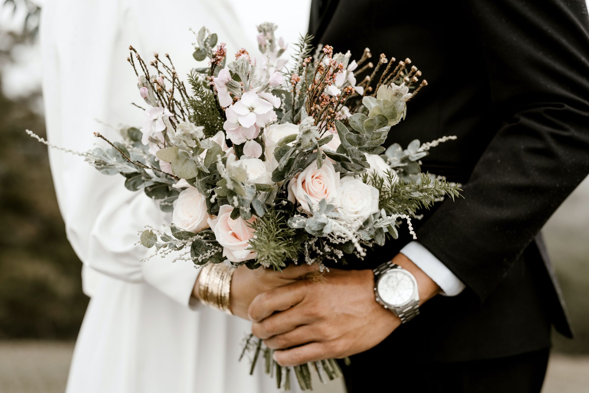 Foto colorida de noivos segurando buquê de flores. A imagem foca nas mãos e no buquê.