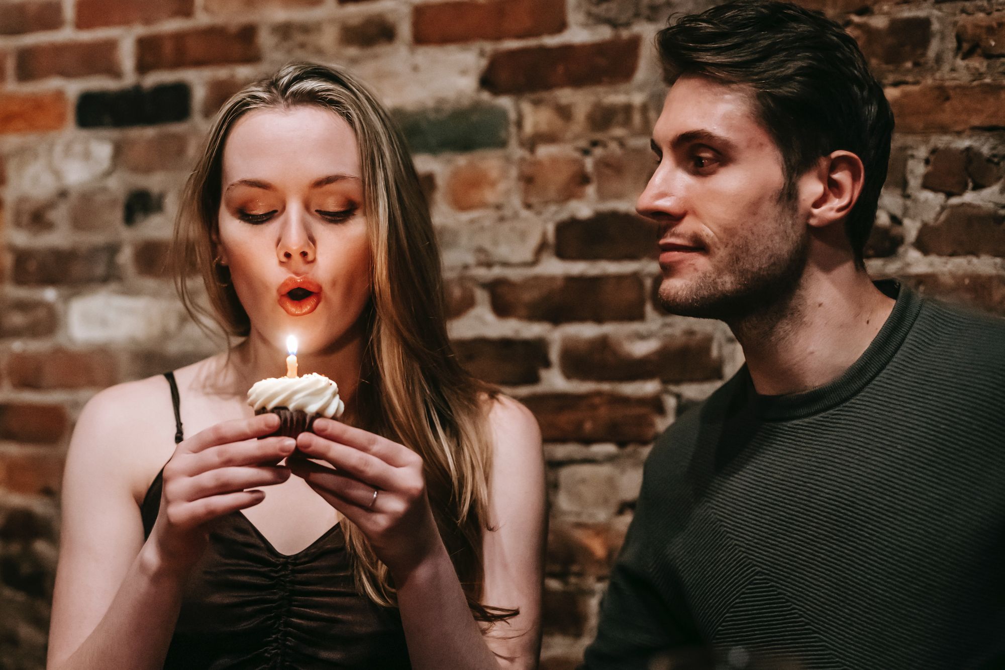 Foto colorida de casal. A noiva está assoprando a vela de um cupcake enquanto o homem olha para ela com olhar apaixonado.