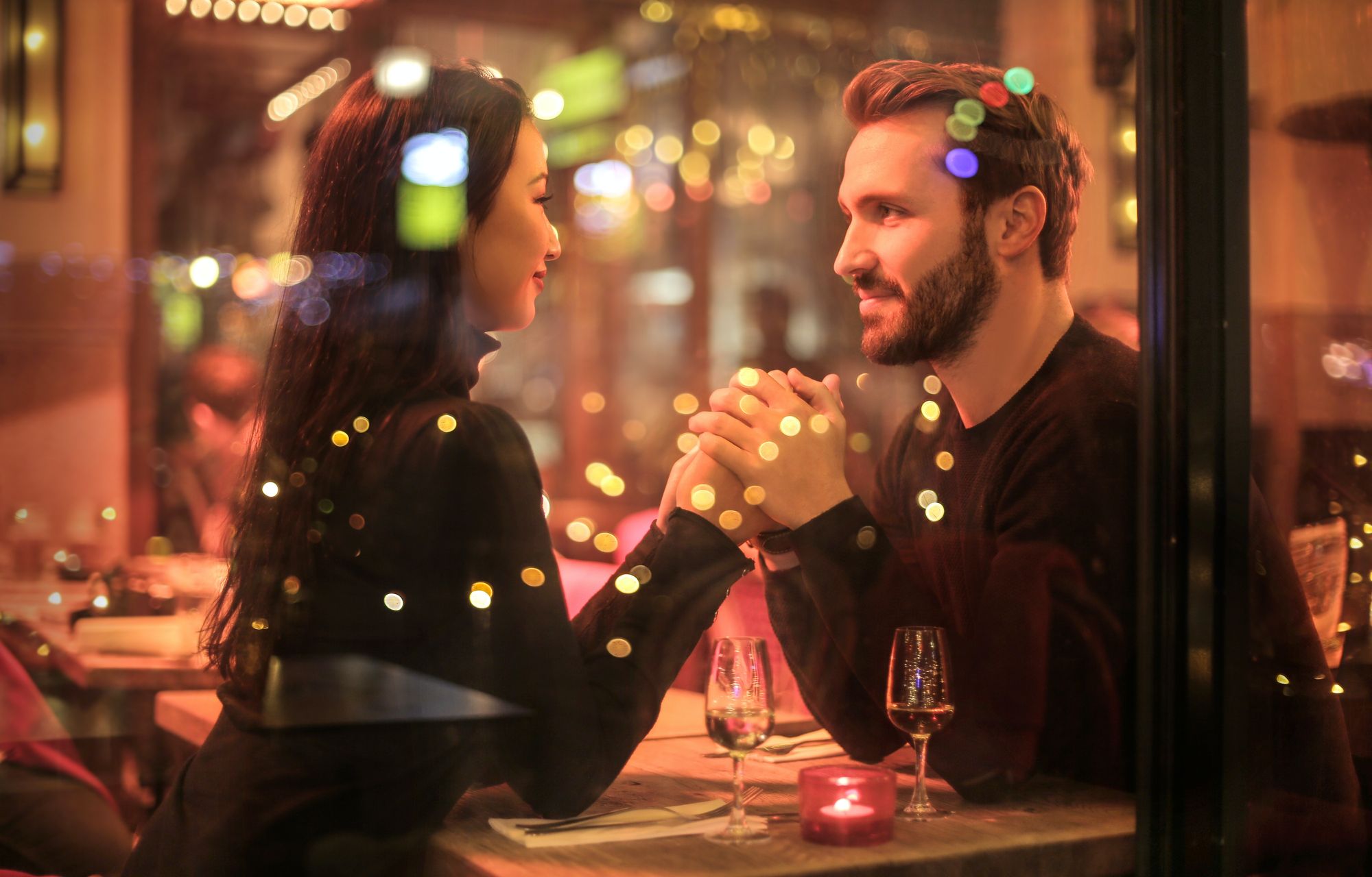 Foto colorida de casal sentados em restaurante. O homem segura as mãos da mulher e olha profundamente em seus olhos.