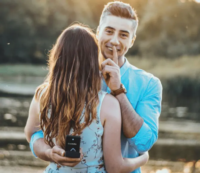 Foto colorida de casal de abraçando. A noiva está de costas para a câmera e o homem está com a caixa de aliança atrás dela, sem que ela veja.