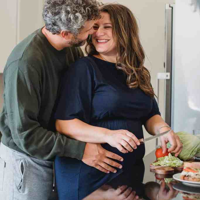 Homem abraçando uma mulher enquanto ela corta a salada, de forma romântica.