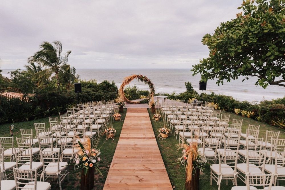 Vista de cima do local e da decoração de um casamento celebrado na praia. As cadeiras são brancas, o tapete é de madeira e há muitas flores enfeitando o caminho.