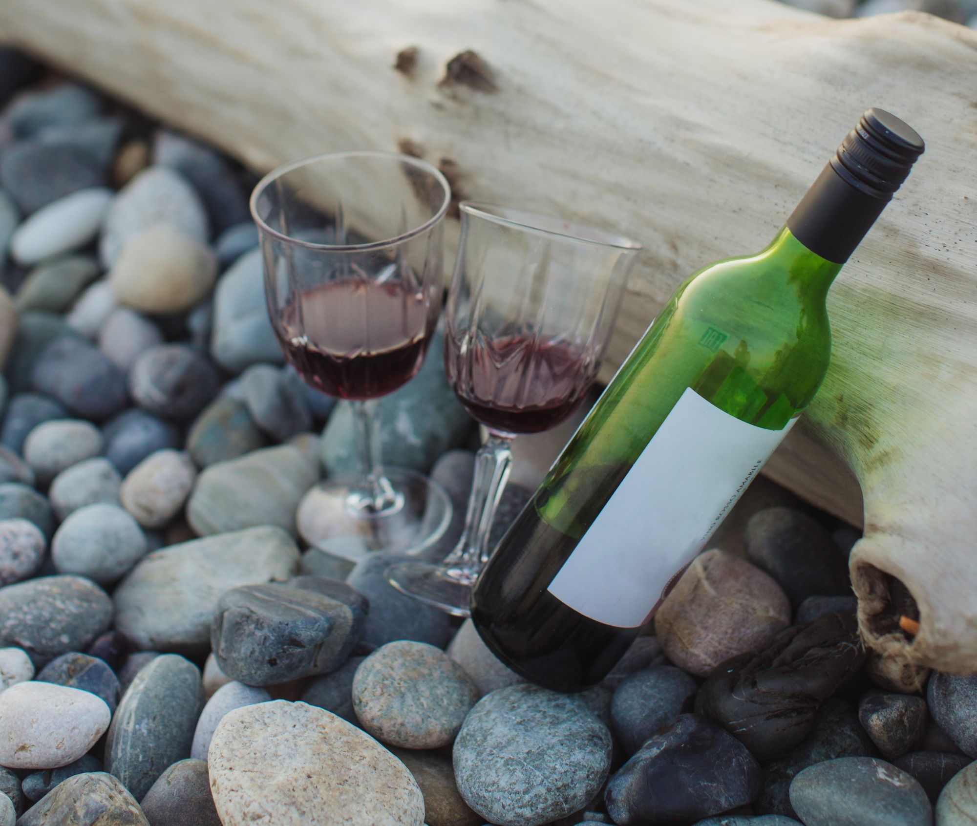 Foto de garrafa de vinho com duas taças ao lado. Os objetos estão escorados num tronco de árvores e apoiados no chão de pedras.
