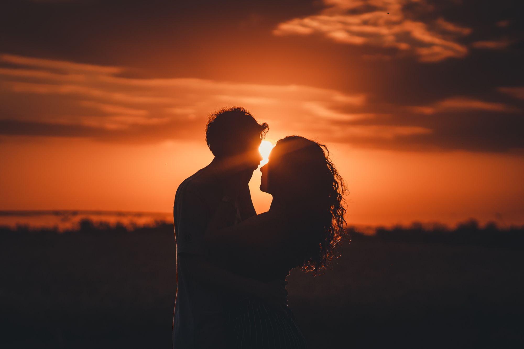 Foto de casal se beijando. Eles estão contra a luz do por de sol.