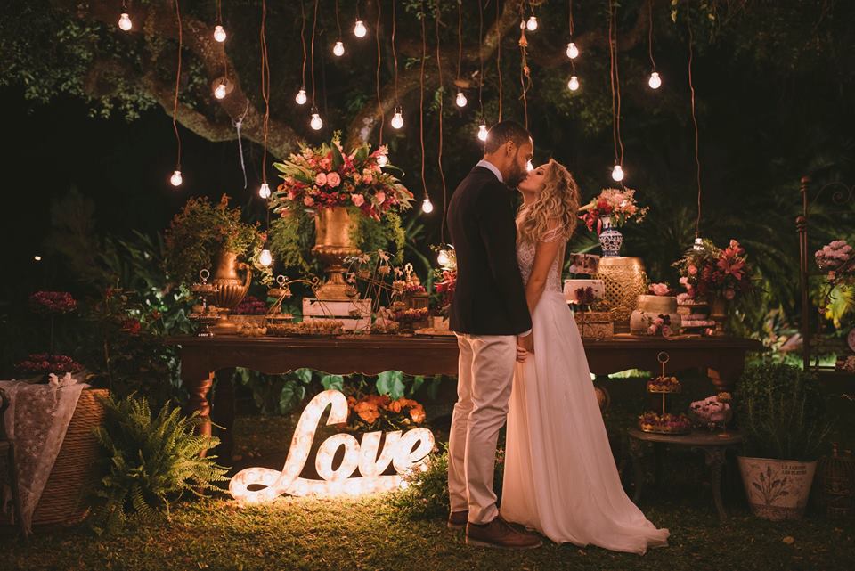 Foto colorida de um casal de noivos se beijando. Eles estão em frente à mesa do bolo.