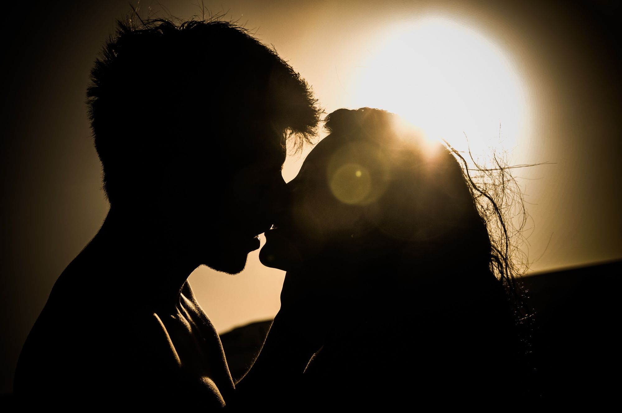 Foto de casal se beijando. Os dois estão contra a luz do sol.