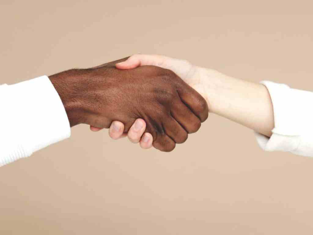 Aperto de mão entre um homem negro e uma mulher branca. Na foto só é possível ver as mãos e que ambos vestem camisa de manga branca.