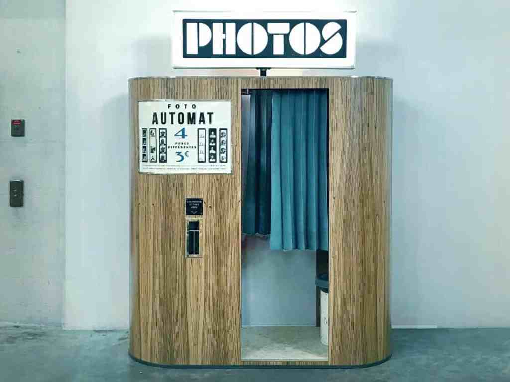 Cabine de fotos de madeira, com cortina azul. Em cima, uma placa escrita "photos".
