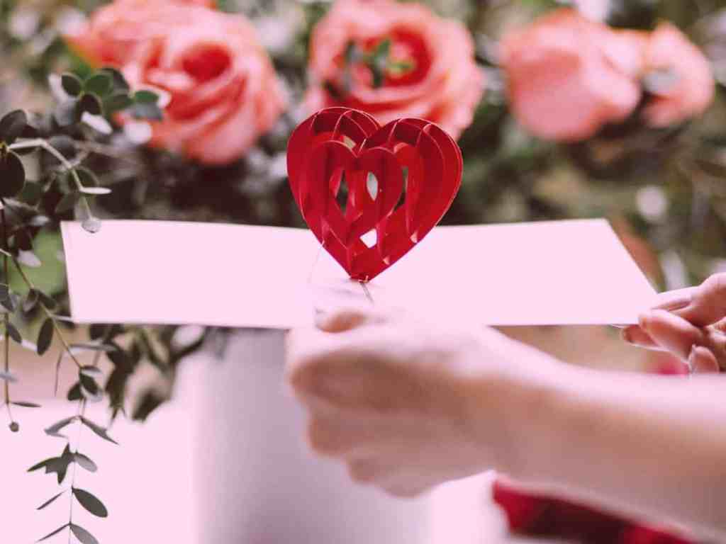 Pessoa segurando um cartão com coração em alto-relevo dentro, no fundo há flores rosas.