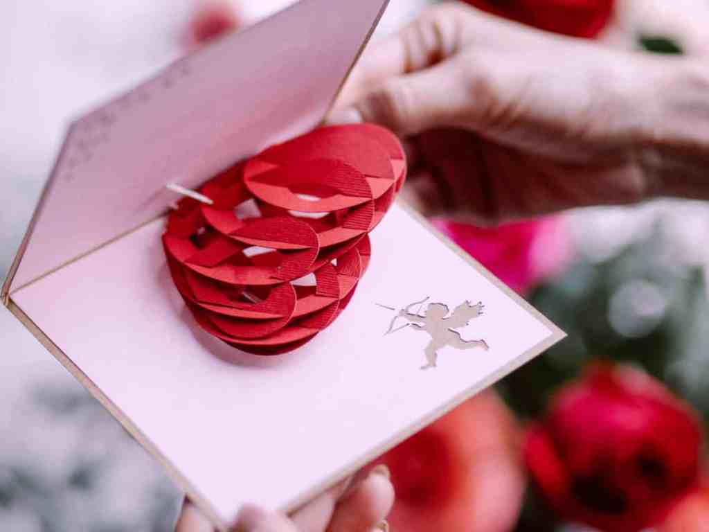 No fundo tem rosas vermelhas desfocadas. Na frente, a mão de uma pessoa abrindo um cartão branco com coração em alto relevo dentro e desenho pequeno do cupido.