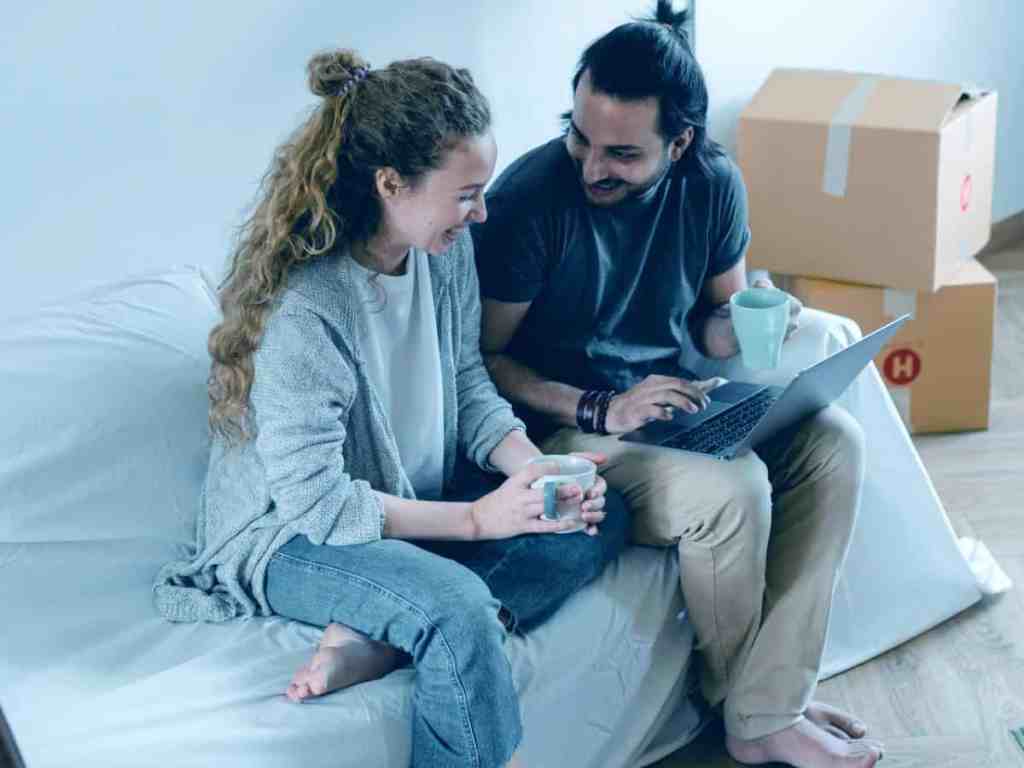 Um homem e uma mulher sentados na cama olhando para tela do notebook e rindo. Ela está de camisa de moletom cinza e calça jeans. Ele está de camiseta verde e calça bege. Ambos tem cabelo longo, o dele é liso e o dela cacheado.
