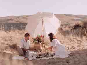 Casal de um homem e uma mulher vestidos de branco. Ambos estão em um campo fazendo piquenique debaixo de um guarda-chuva branco, com uma cesta de flores atrás. Esta é uma ideia bem romântica para o dia dos namorados.