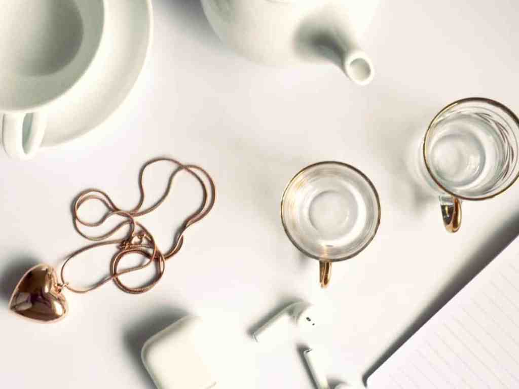Mesa branca, em cima dela têm: com xícaras, bule, fones de ouvido e um colar dourado em forma de coração