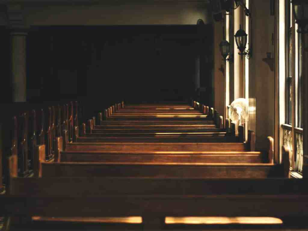 Imagem de dentro de uma igreja vazia. É uma fileira de bancos.