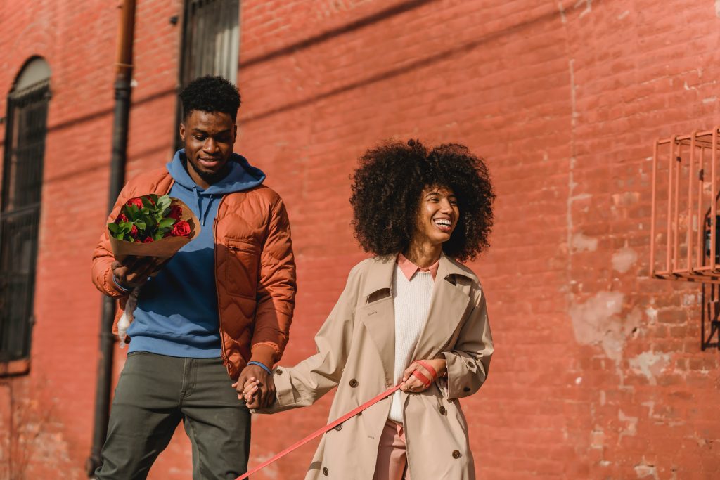 Foto de casal caminhando por rua. O homem segura um buquê de flores.