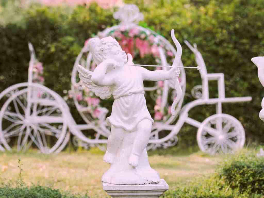 Estátua do cupido com arco e flecha em um jardim, de fundo há uma estátua de carruagem com flores de ornamento.