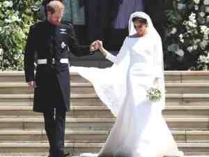 Megan markle e principe harry nas escadas no dia do casamento, ele usa sobretudo e calça preta e ela usa um vestido de casamento branco com decote ombro a ombro. Inspire-se neste vestido de casamento real britânico.