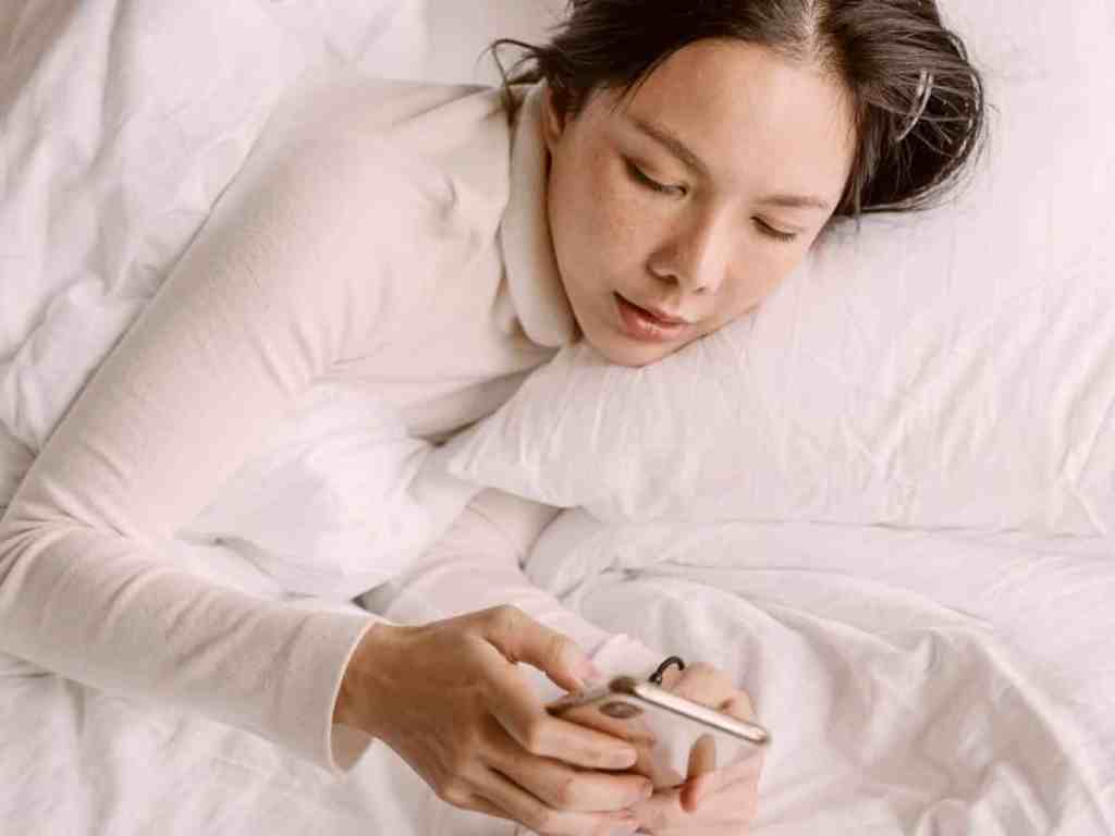 Mulher de blusa longa branca, com cabelo castanho escuro bagunaçado repartido no meio, está deitada na cama mexendo no celular iphone. Os lençois, o travesseiro e a capa do celular são brancos também.