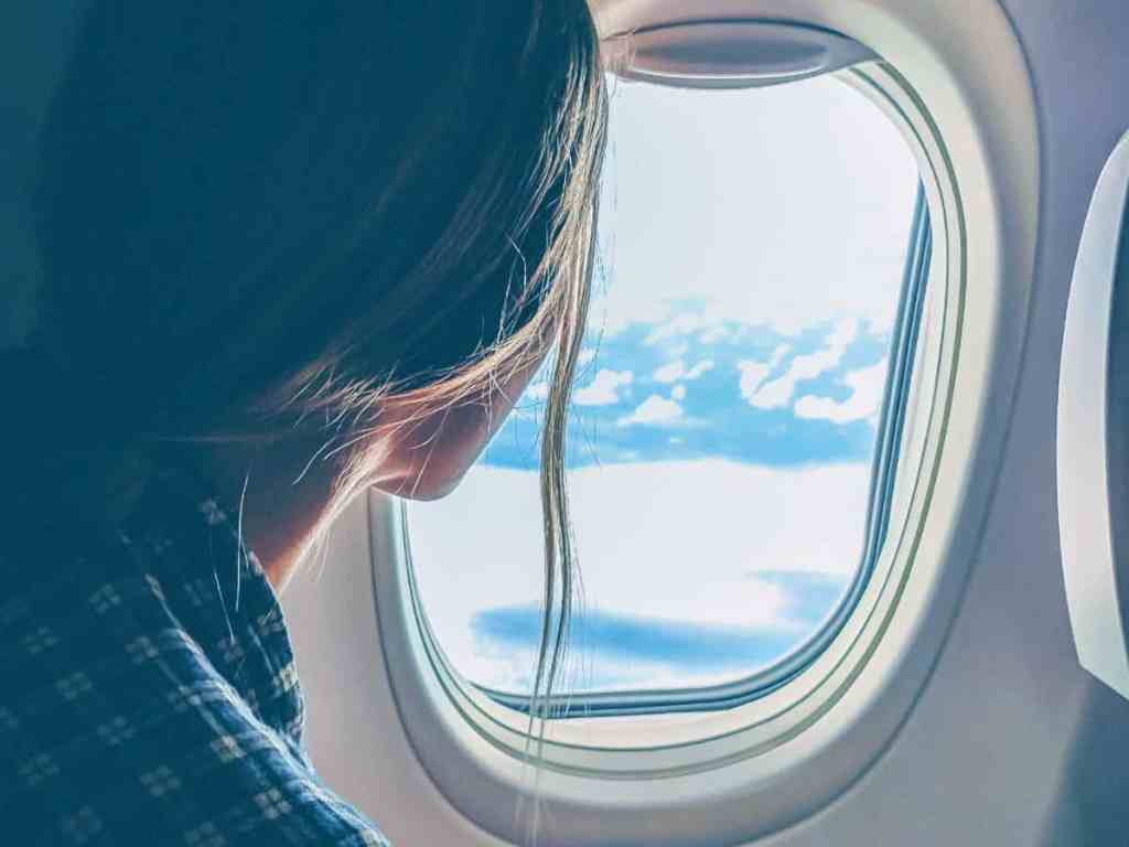Mulher vestindo camisa xadrez azul e cabelo meio preso, olhando pela janela do avião, o céu está azul claro.