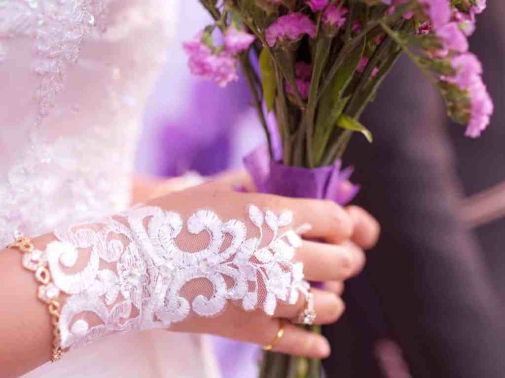 Noiva de vestido branco, na mão uma luva aberta de renda, pulseira dourada e anel dourado no dedo mindinho, ela segura um buquê de flores lilás;- imagem do post "quais acessórios usar no casamento? "