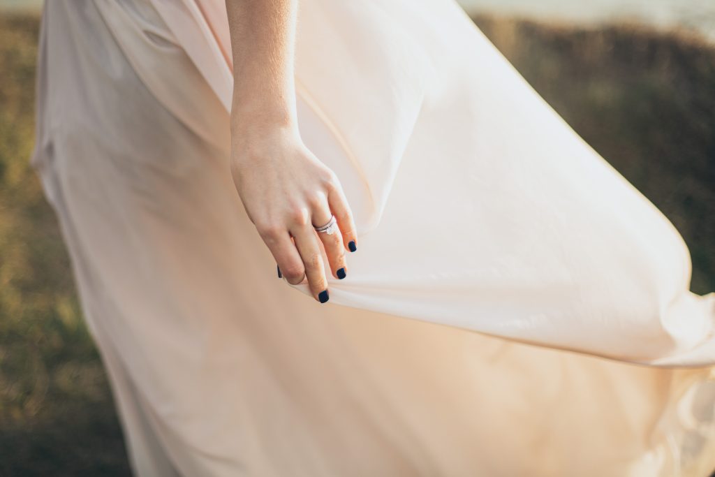 Foto de mulher segurando vestido com a mão. Ela está usando dois anéis: um de casamento e outro de noivado.