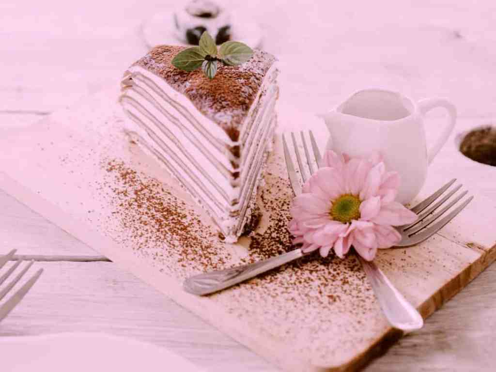 Torta servida em cima de uma bandeja de madeira. Junto têm dois garfos, em cima deles uma flor da cor rosa, atrás um bulê branco pequeno.