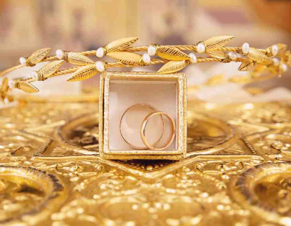 Qual a diferença entre quilates e kilates? - Aliancas-caixa-ouro-tiara-1 Caixinha de ouro com duas alianças dentro. Está em cima de uma mesa de ouro com vários desenhos e em cima da caxinha tem uma tiara douada com detalhes brancos.