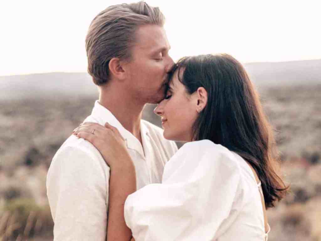 Primeiro ano de casamento: problemas comuns e como passar por eles - Casal-abracados-abraco-beijo-na-testa Casal abraçado. Ele tem cabelo castanho claro liso penteado para trás e ela tem cabelo preto liso e de franja. Ambos estão usando branco.