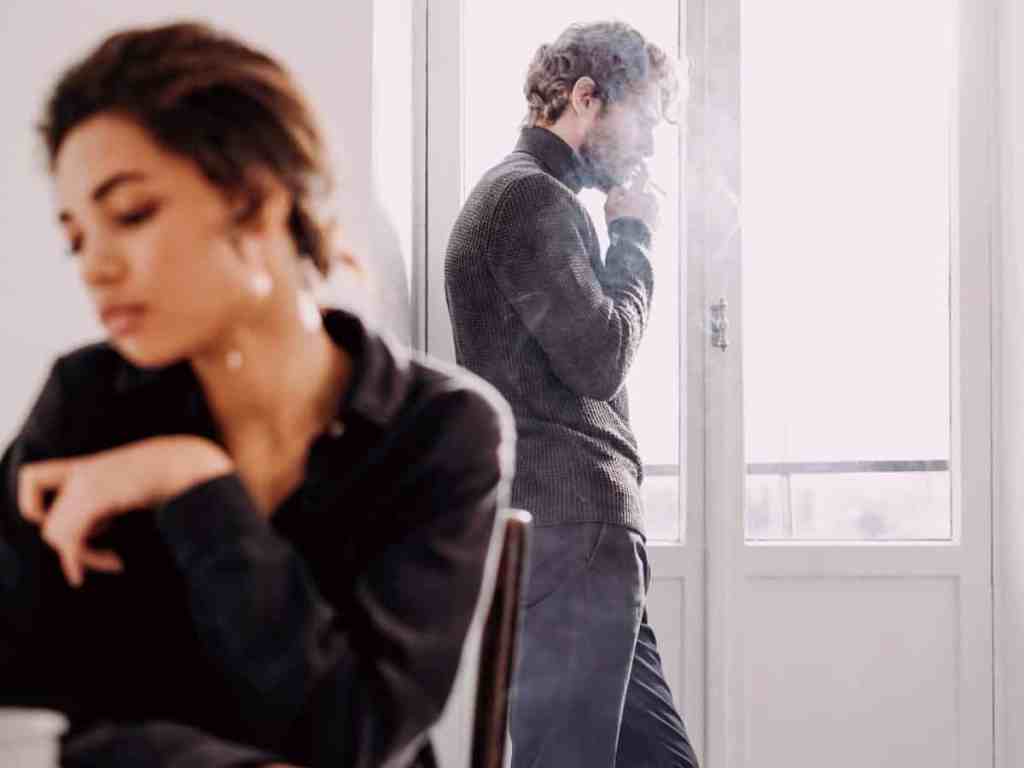 Primeiro ano de casamento: problemas comuns e como passar por eles - Casal-briga-brigados-fumando Casal distante brigado. Ela está sentada numa cadeira com o rosto triste, ela está com cabelo preso e jaqueta. Já ele está fumando perto da janela e usa roupa todda preta