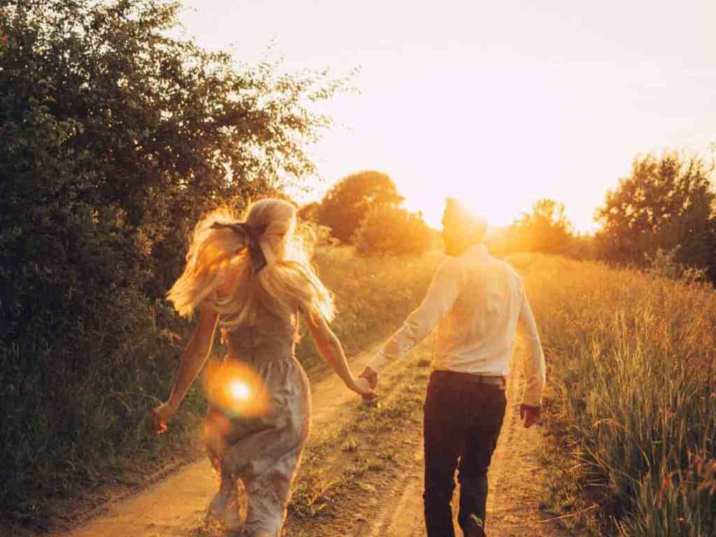 Casal andando de mãos dadas no campo ao por do sol. Ela está de vestido e cabelo loiro solto, ele está de camisa branca e calça preta.