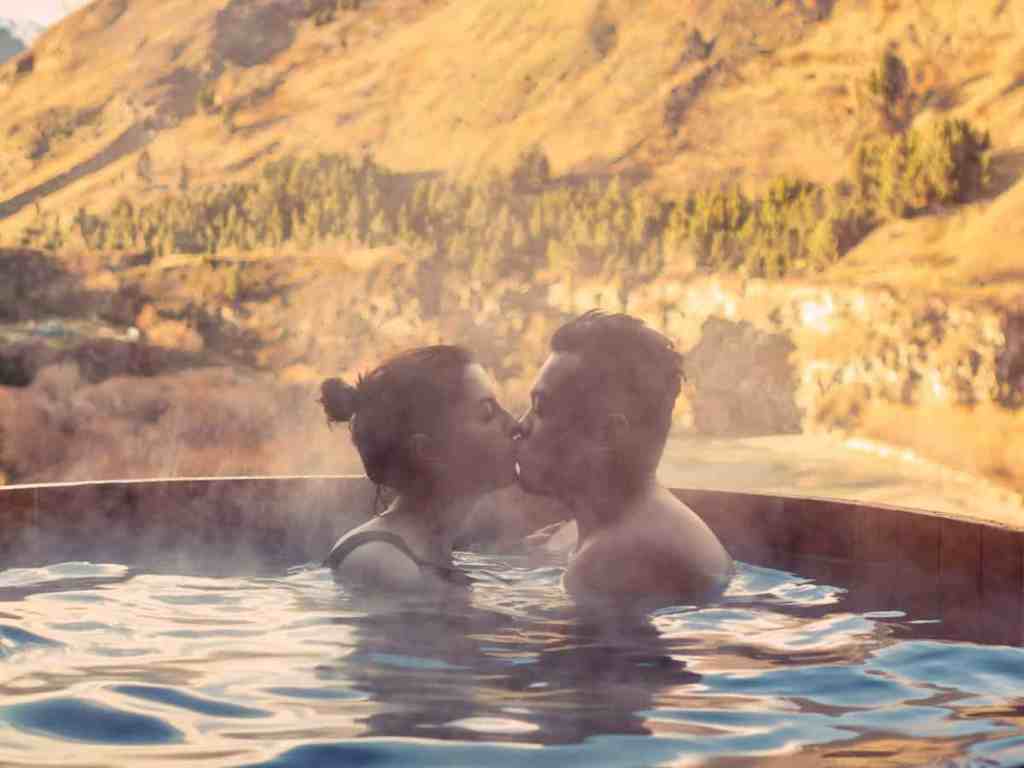 Casal dentro de uma piscina redonda se beijando. De fundo há uma paisagem verde, é um local mais isolado.