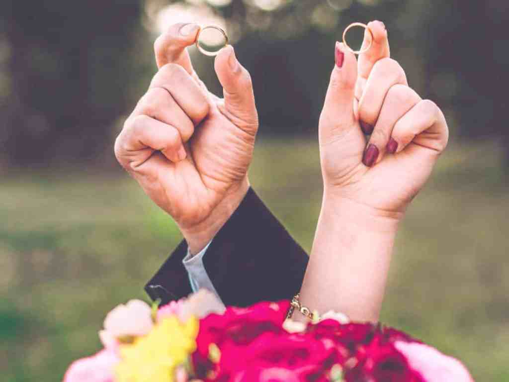 Foto focada na mão do casal, cada um segura para cima sua aliança de ouro, na frente tem um buquê de flores.