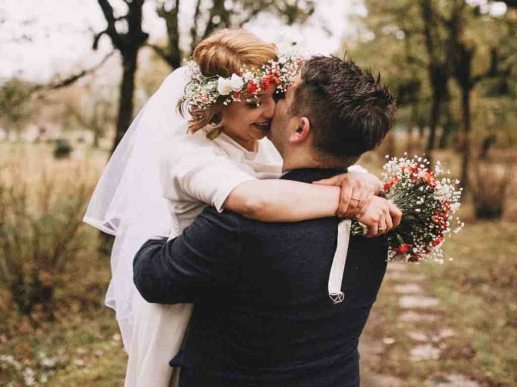 Noivos se beijando abraçados. Ela está de vestido de casamento, véu, tiara de flores e flores na mão. Enquanto ele veste terno preto.