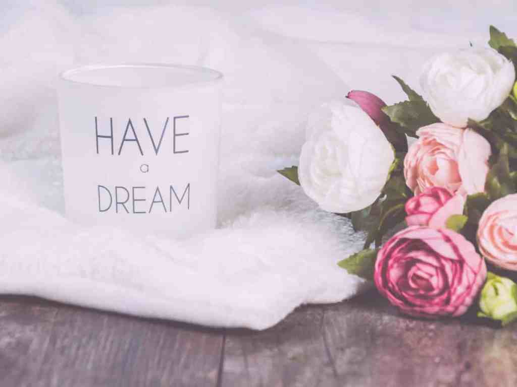 Caneca escrito "have a dream", ou seja, "tenha um sonho", em português, junto com um buquê de flores rosas e brancas no chão de madeira sob um vestido de noiva branco.