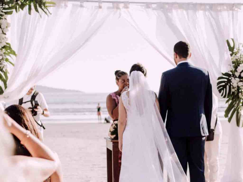 Noivos casando na praia, atrás tem vários convidados sentados.