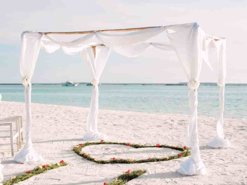 Orla da praia com tenda aberta branca e coração no chão feito de folhas.